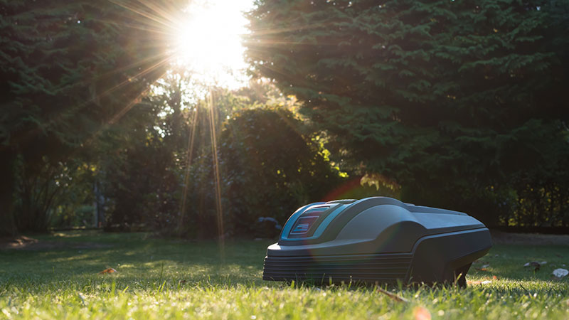 Een robotmaaier staat op een grasveld met op de achtergrond het zonnetje dat door de bomen schijnt